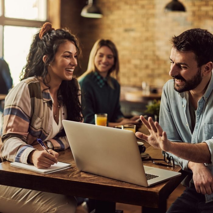 Happy man using laptop and talking to his female friend who is taking notes in a cafe.